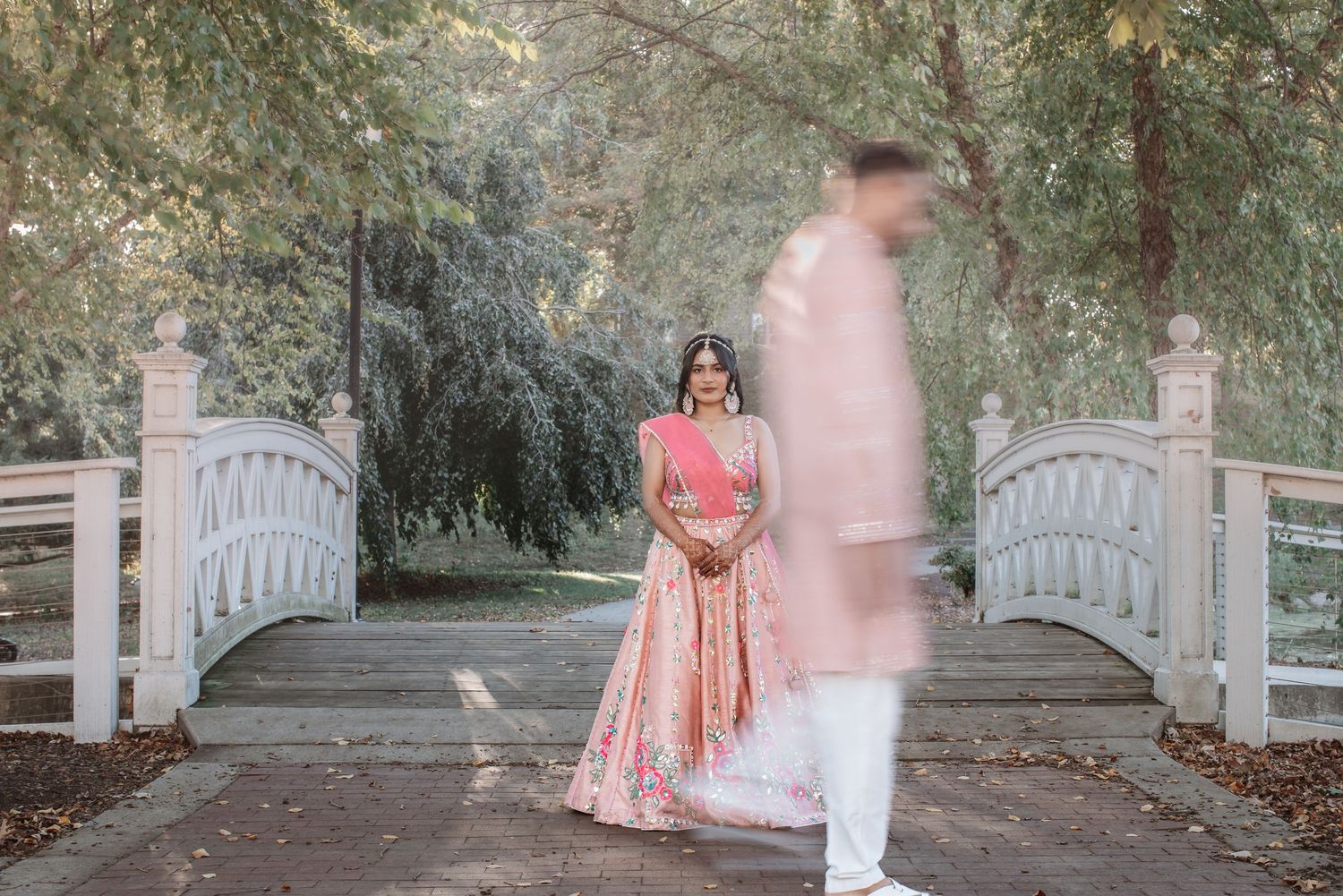 Indian bride in pink lehenga stands on bridge as groom walks past in blur.