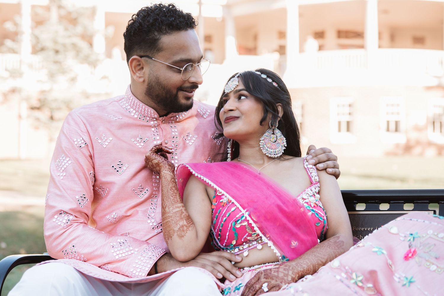 Indian couple in matching pink outfits smiling at each other during pre-wedding mehendi ceremony.