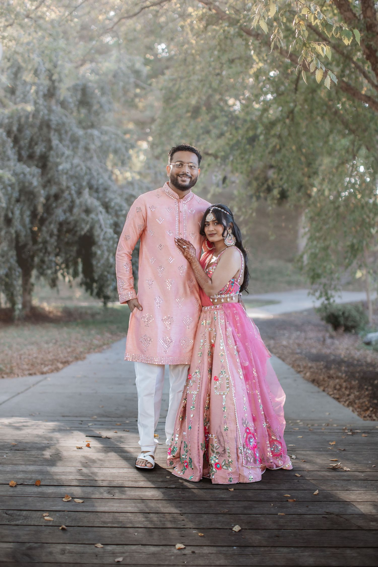 Indian couple in coordinating pink traditional attire posing on wooden path in park.