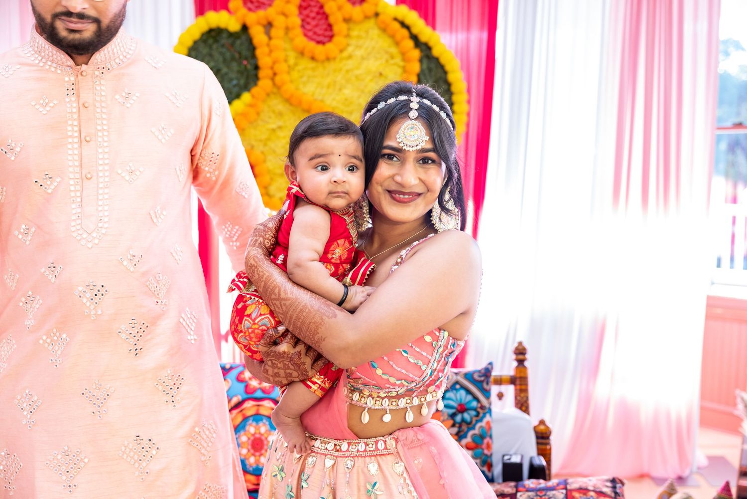 Indian bride in red lehenga holding baby at traditional mehendi wedding ceremony.