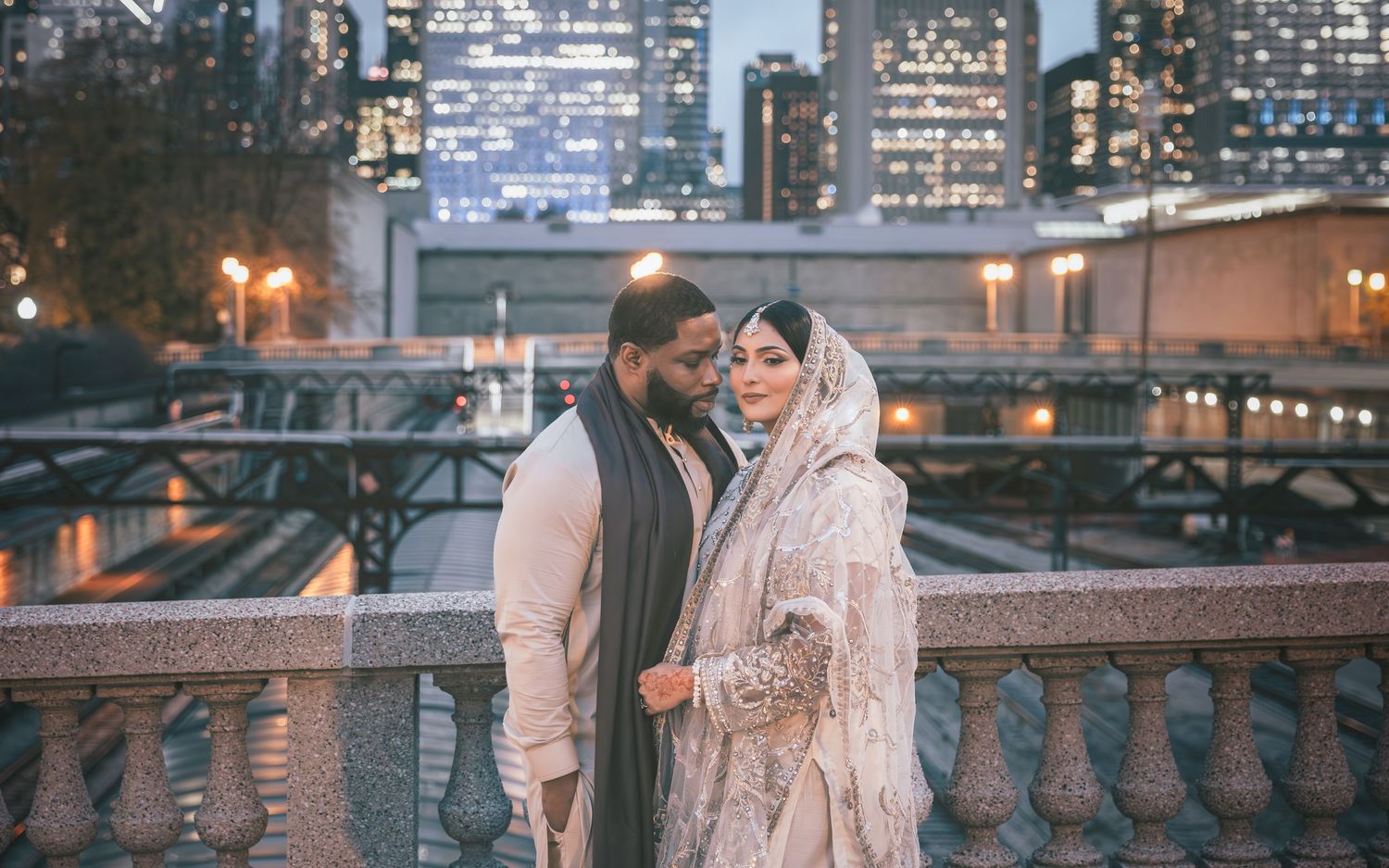 Groom in gray shawl and bride in embroidered lehenga share intimate moment with city skyline backdrop.