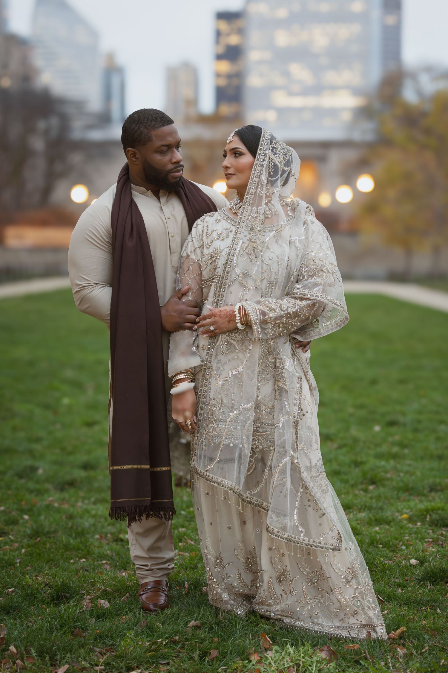 Interracial wedding couple in traditional attire pose elegantly with city lights glowing in the background.
