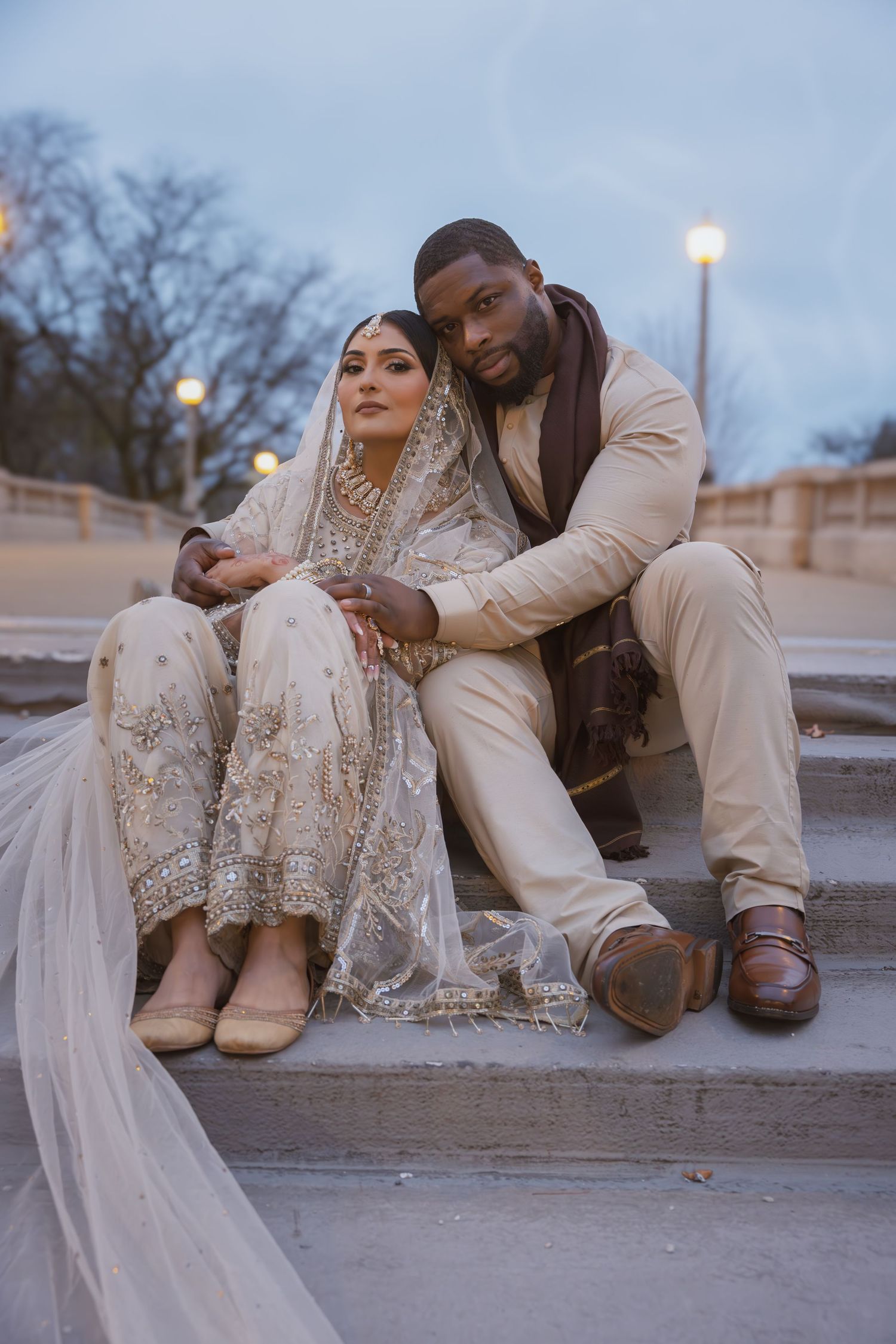 South Asian bride in ivory embroidered lehenga with long veil sits with groom on outdoor steps.