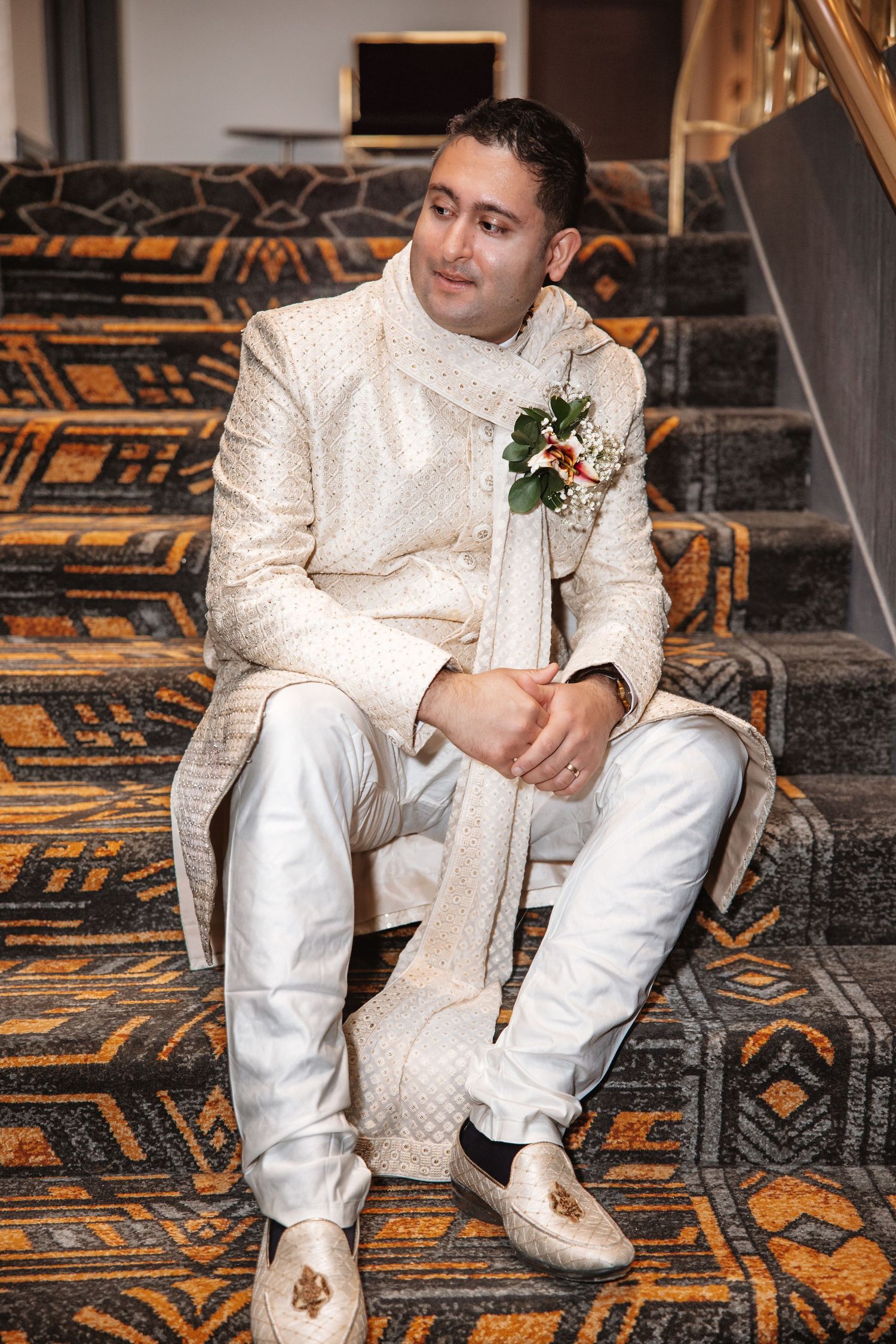 Groom in cream embroidered sherwani with boutonniere sits on ornate carpeted stairs.