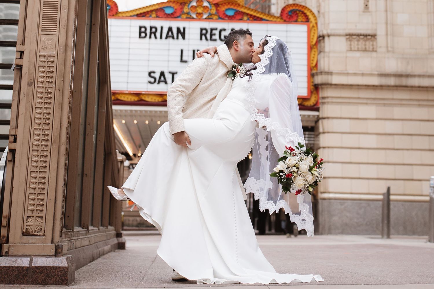 Groom lifts and kisses bride in white gown outside Chicago Theatre marquee on wedding day.
