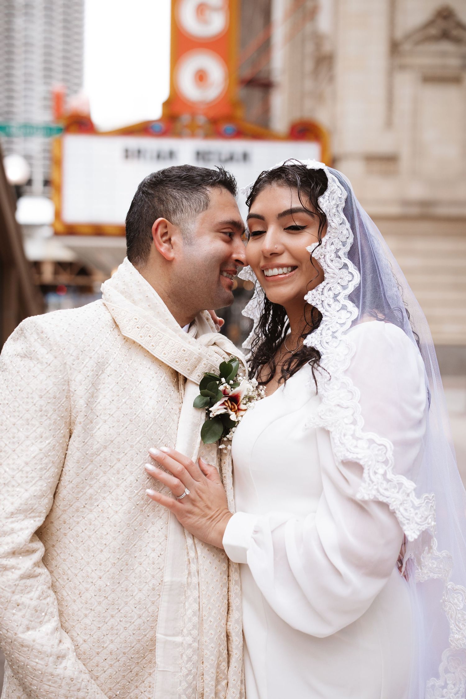 Smiling South Asian bride in lace veil and groom in cream sherwani embrace outside Chicago Theatre.