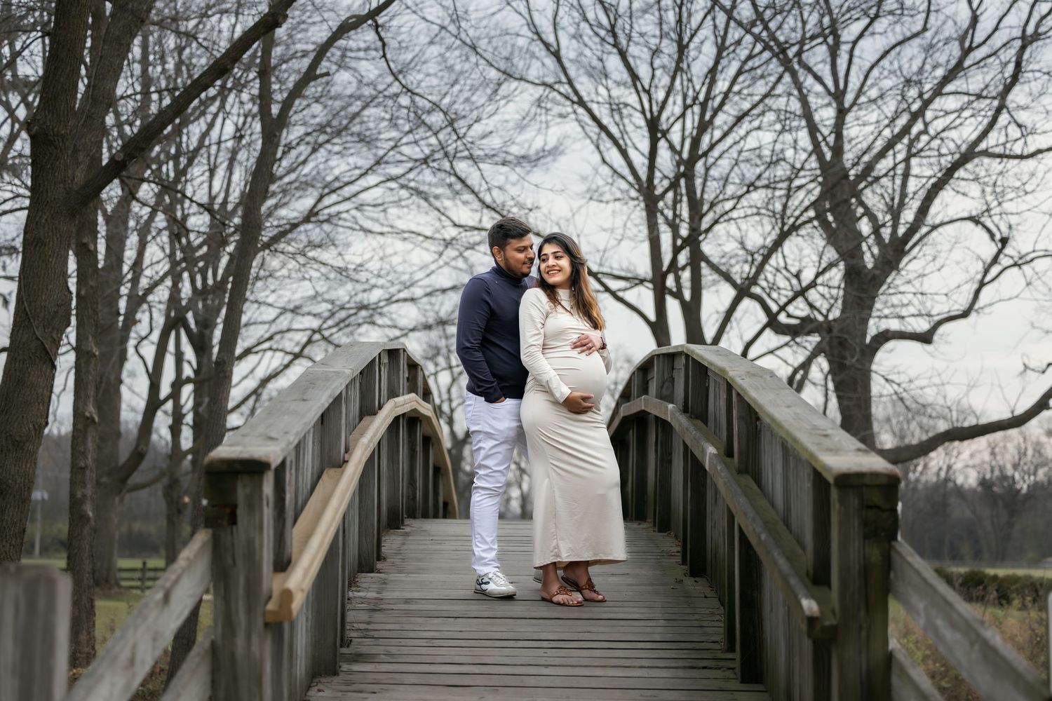 Loving pregnant couple embracing on a wooden bridge surrounded by bare trees.