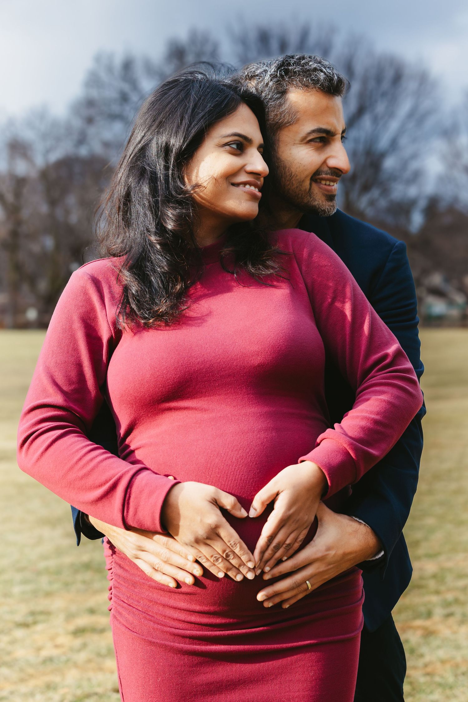 Smiling pregnant couple outdoors in park, both forming heart shape with hands on her baby bump.