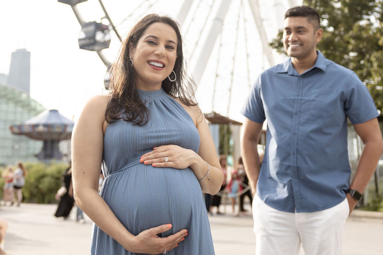 Smiling pregnant woman in blue dress holding belly near ferris wheel with partner behind her.