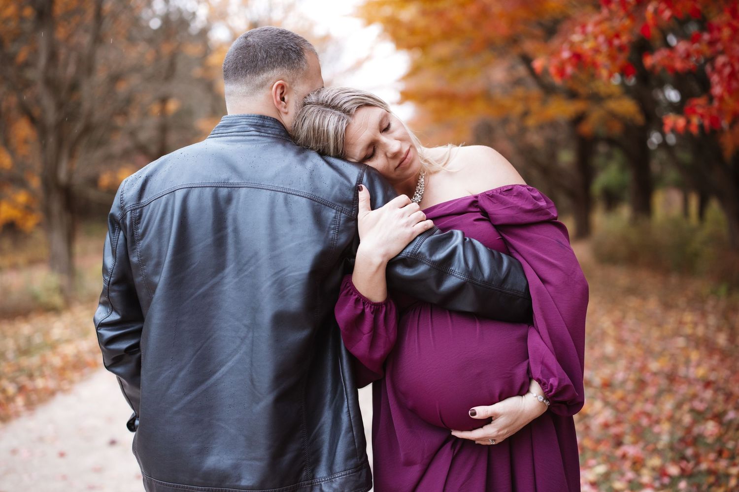 Pregnant woman in burgundy off-shoulder gown embraces man in black leather jacket on fall foliage path.