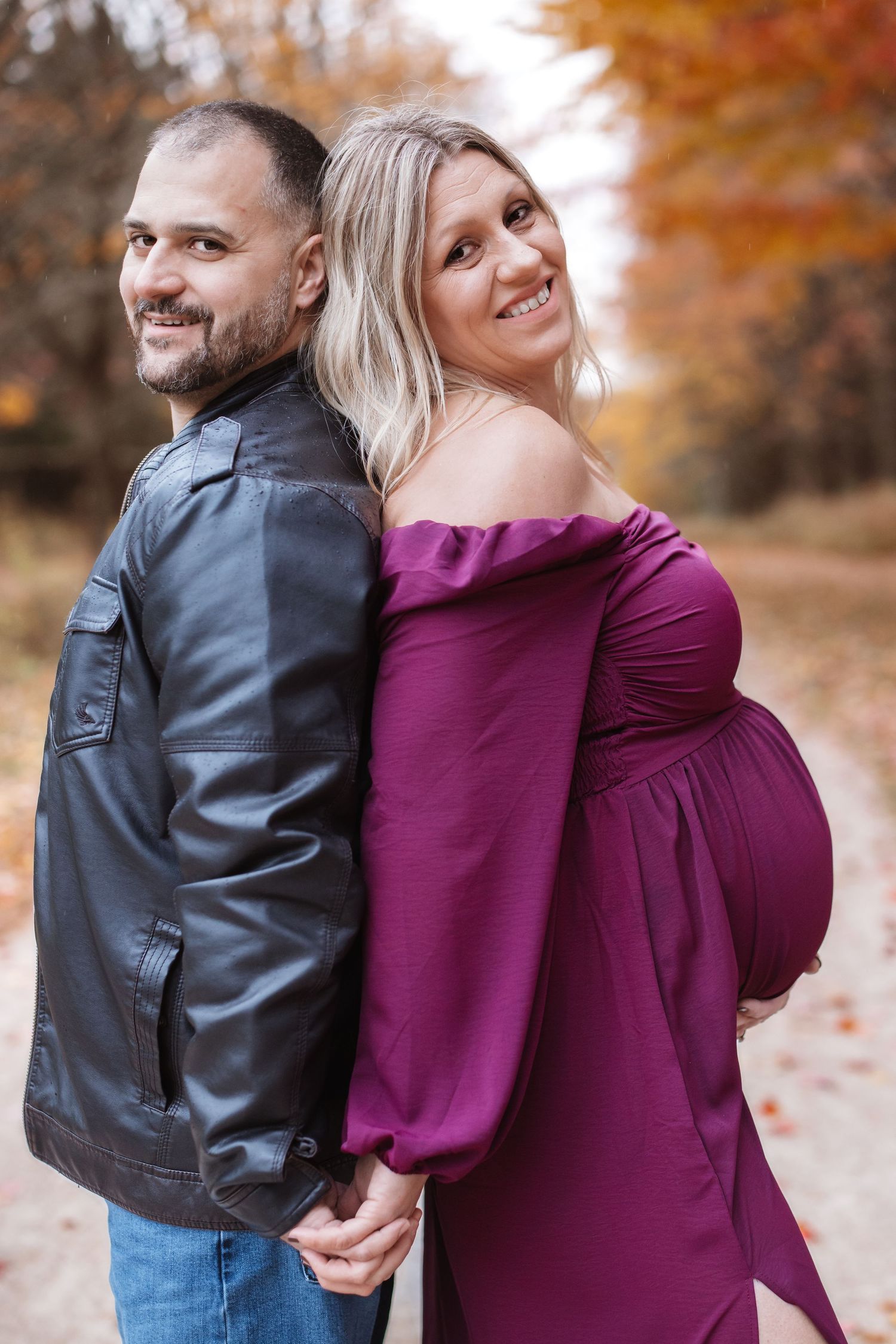 Smiling expectant couple stands back-to-back holding hands on autumn trail, woman in purple maternity gown and man in leather jacket.