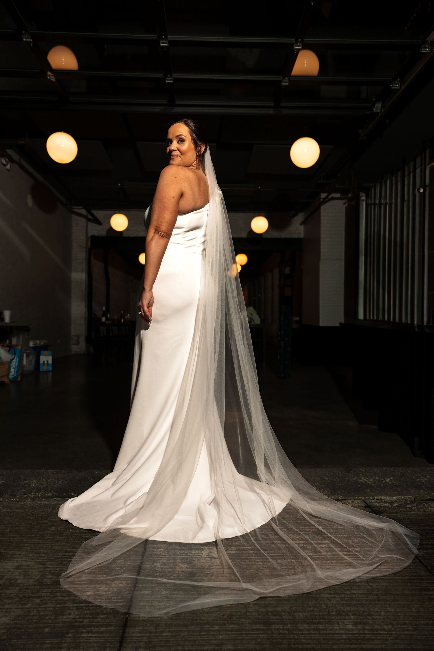 Bride in sleek white one-shoulder gown with long cathedral veil turns gracefully showing back in atmospheric venue.