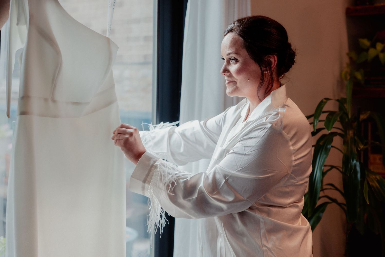 Smiling bride in white robe touching her wedding dress hanging by a sunlit window.
