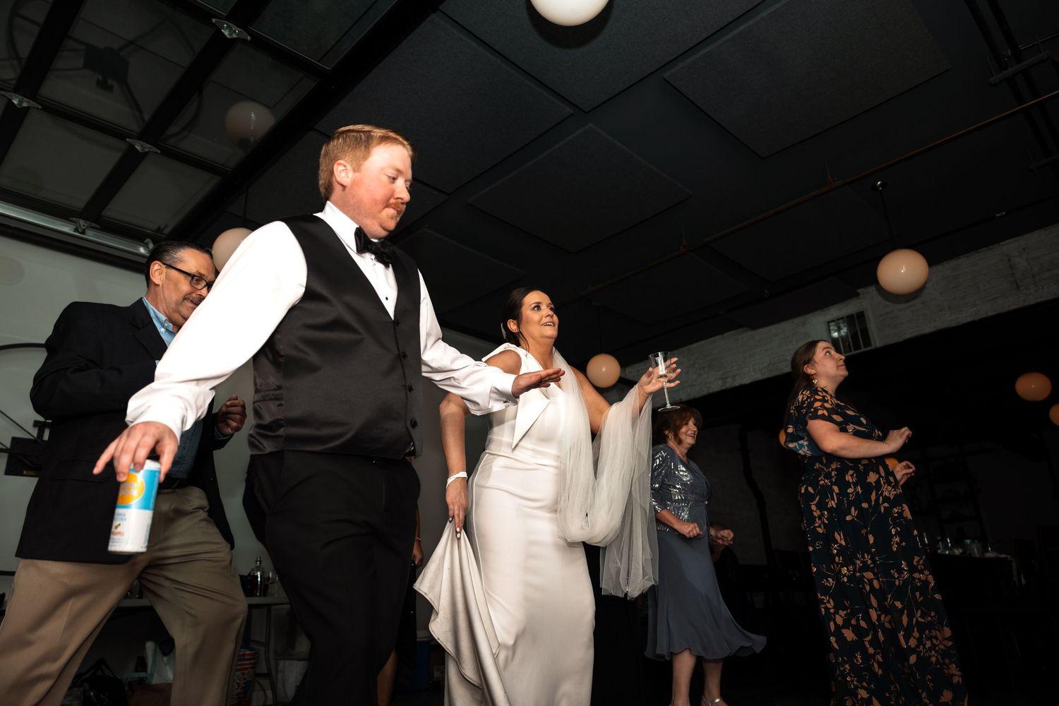 Bride in white gown dances energetically with guests in black vests at lively wedding reception.