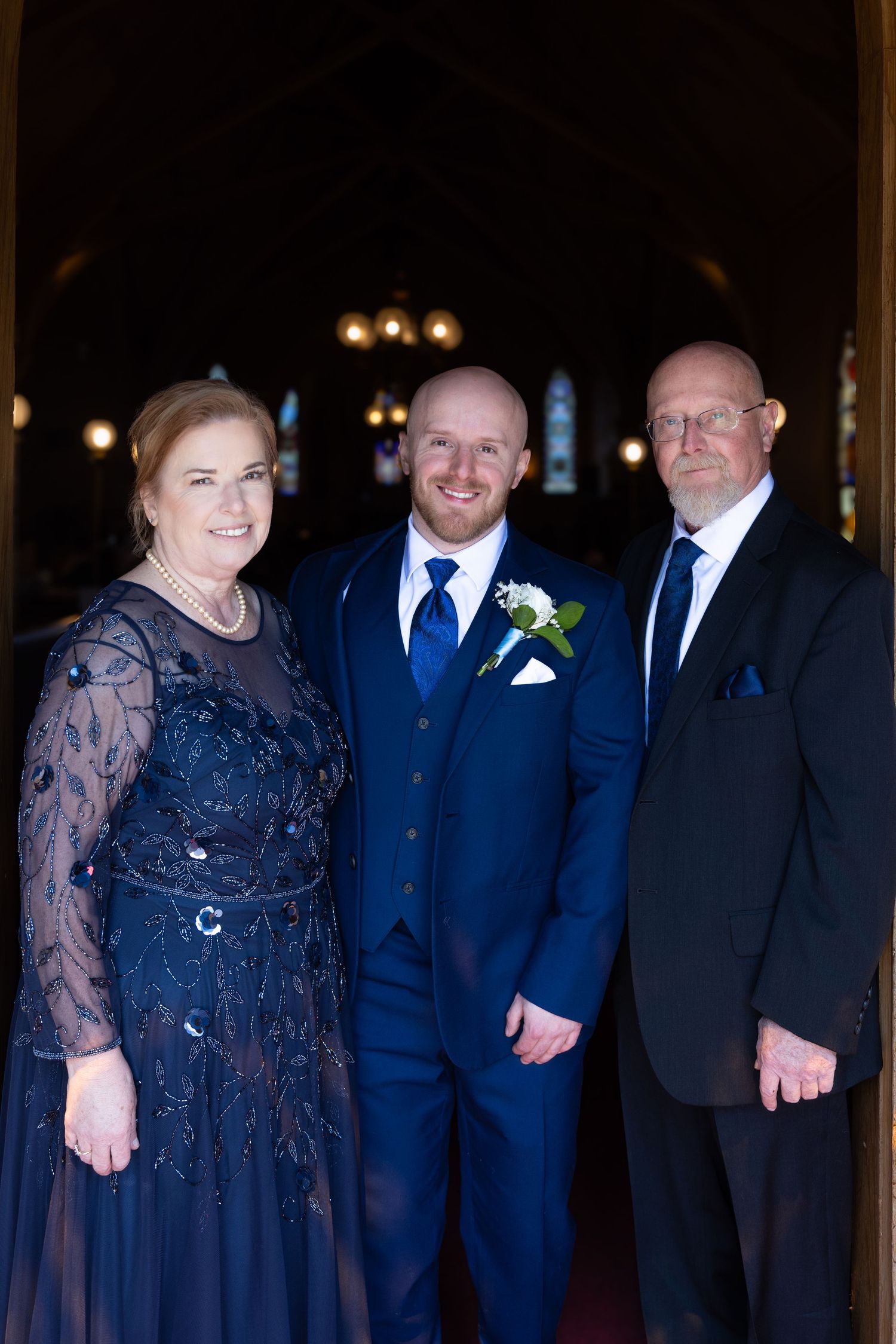 Groom in navy blue suit smiles with parents at church entrance before wedding ceremony.