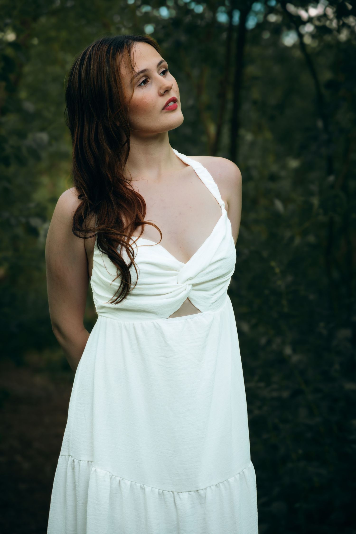 A brunette woman in a white halter dress gazes upward while standing among lush green trees outdoors.