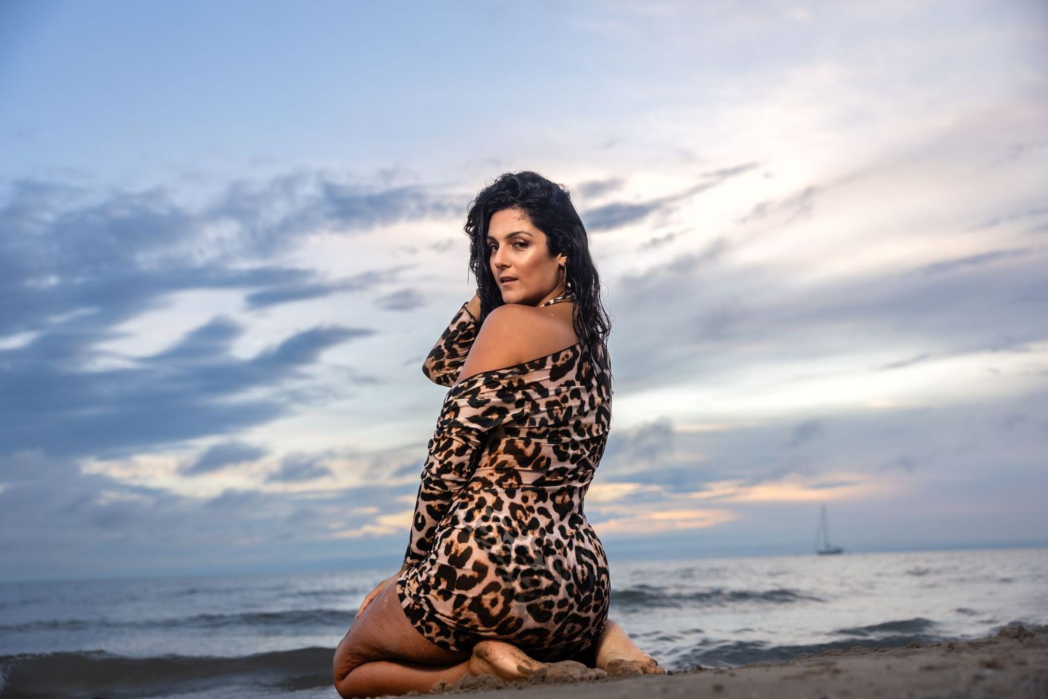 Woman in leopard print dress kneeling on beach at dramatic sunset.