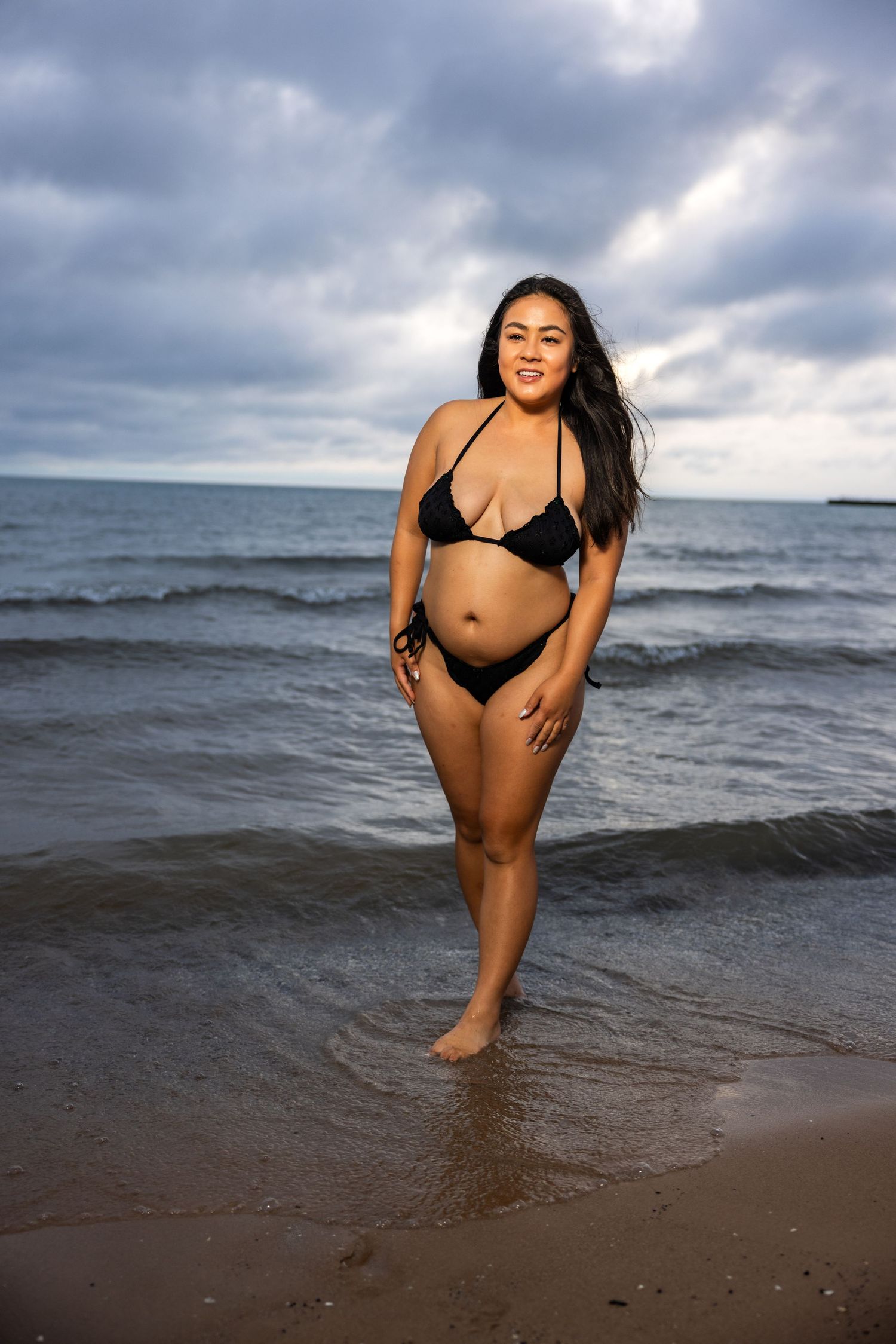 Smiling woman in black bikini standing on sandy beach shoreline with ocean waves and cloudy sky.