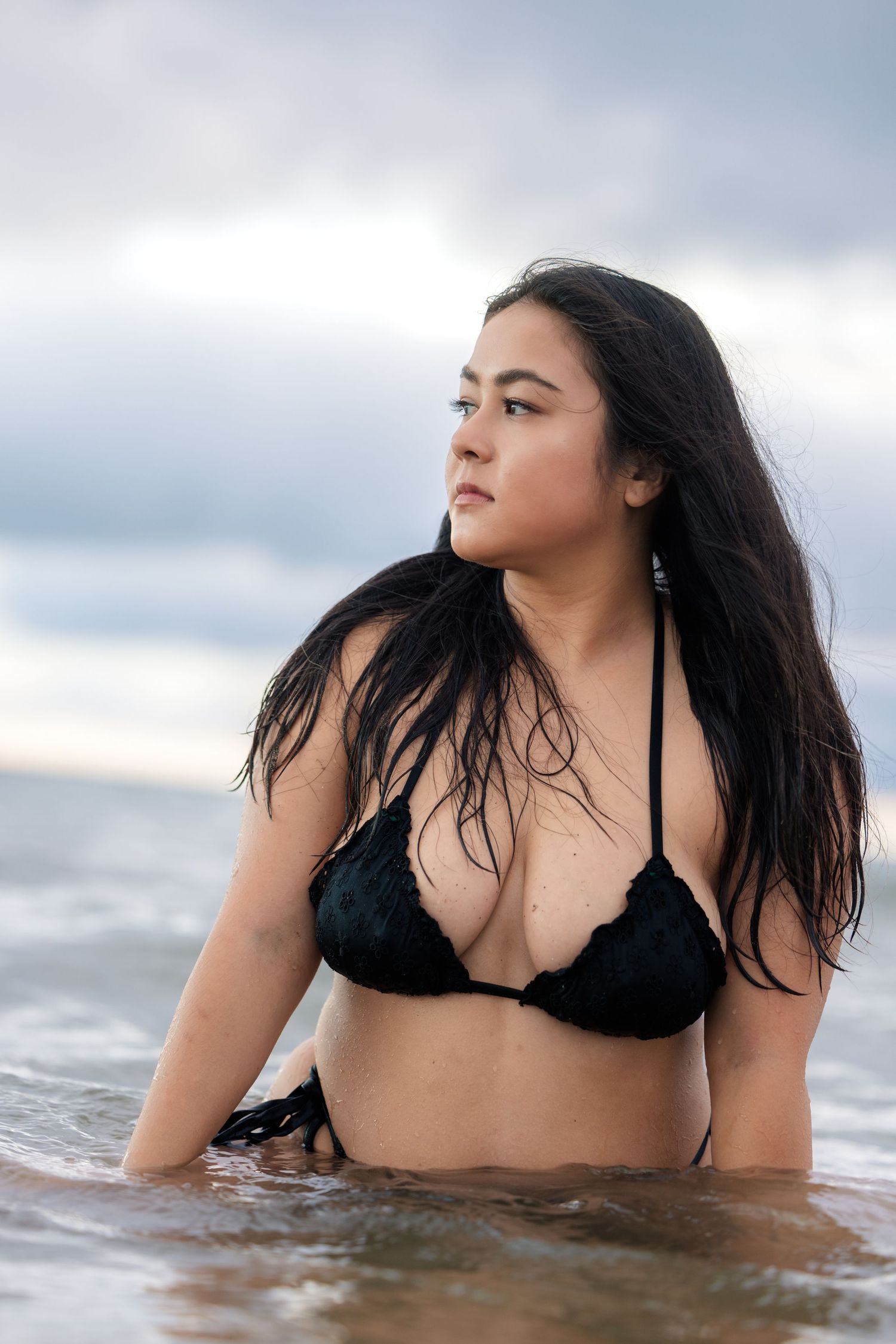 Plus size woman in black bikini kneeling in ocean surf with long dark hair on cloudy day.