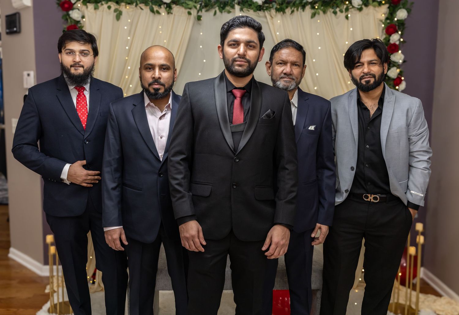 Five men in formal attire pose together at a wedding venue adorned with red and white floral arrangements.