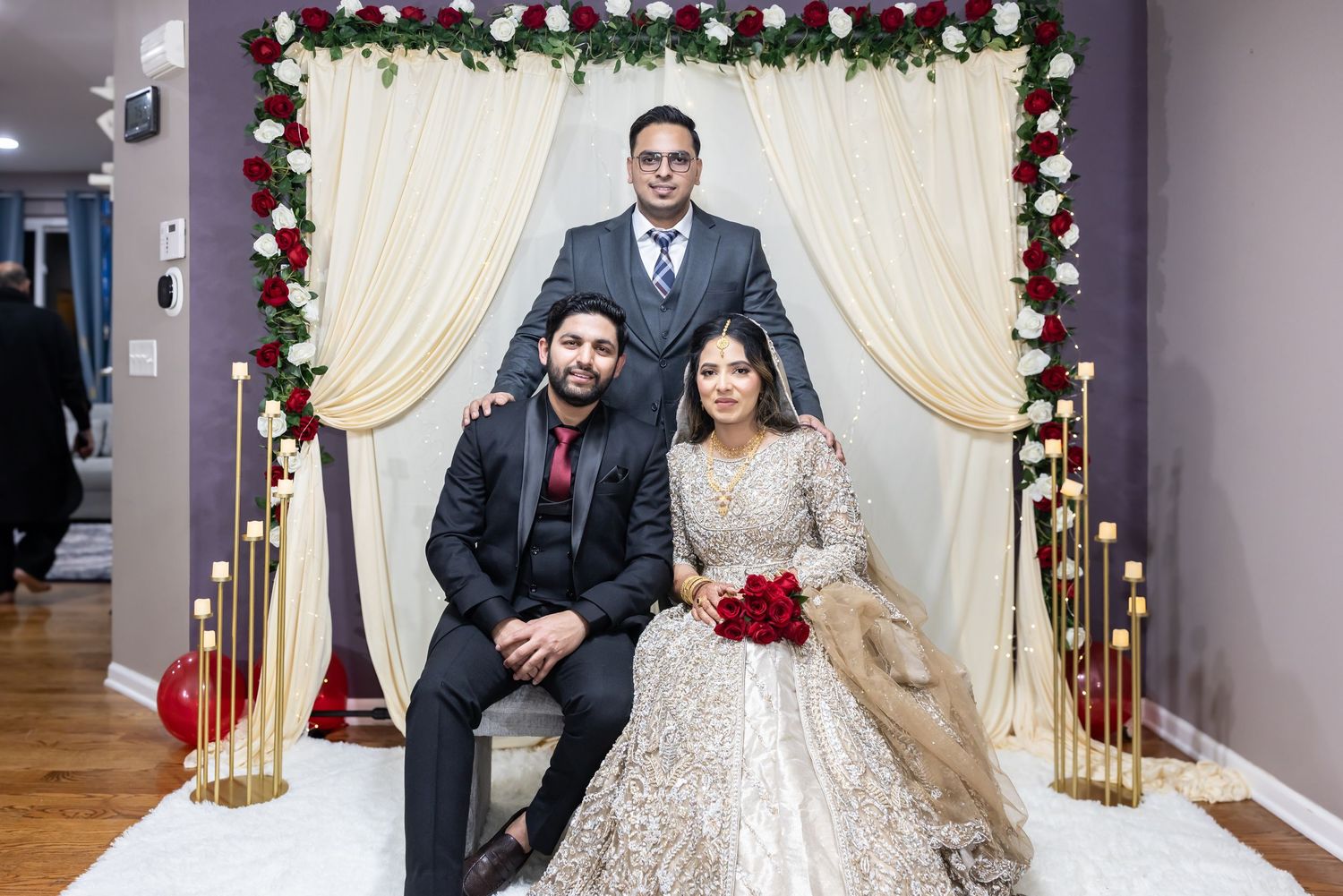 Bride in gold lehenga poses with groom in black suit and elder man in gray suit at decorated wedding backdrop.
