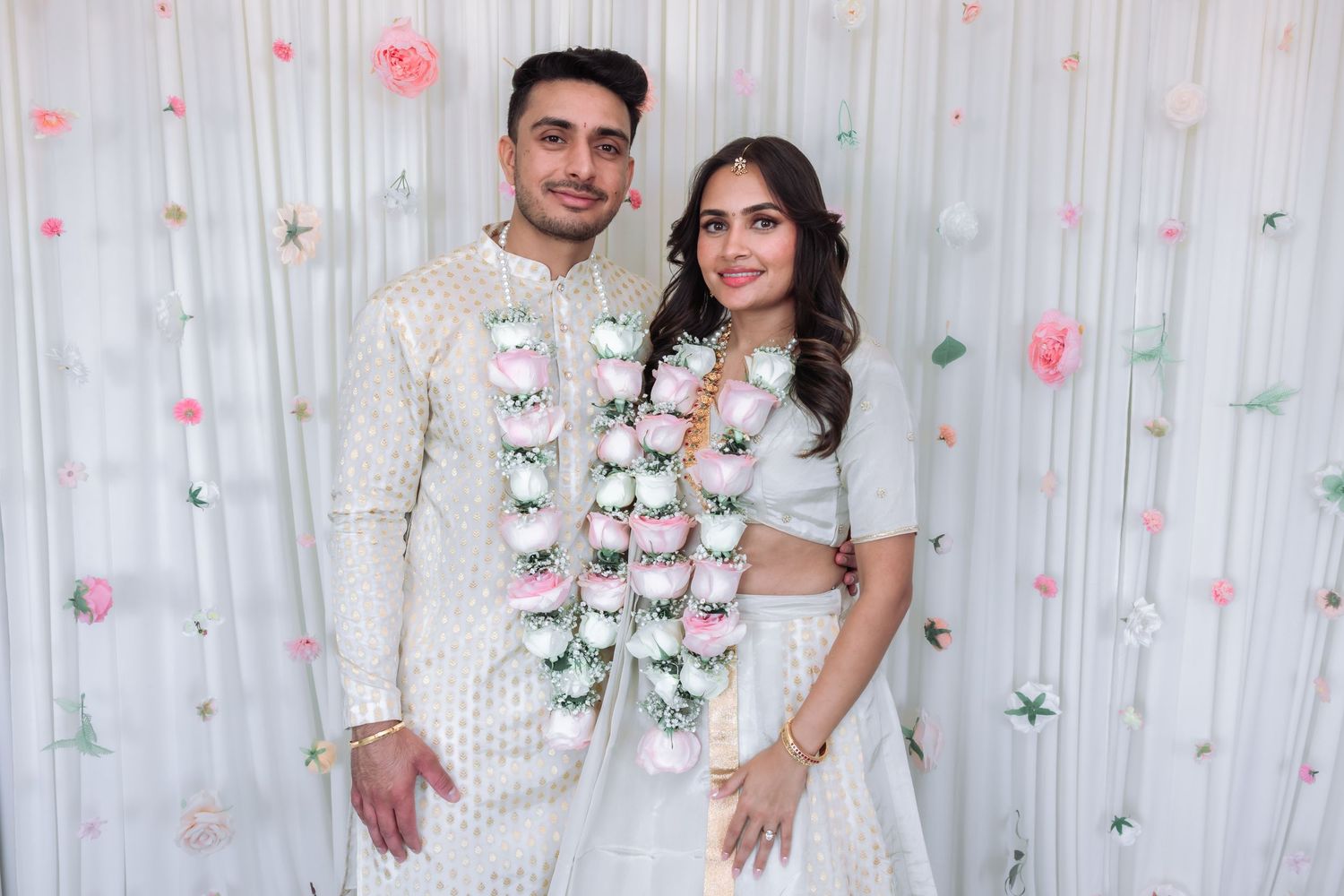 Indian couple in matching white outfits exchange floral garlands during traditional engagement ceremony.