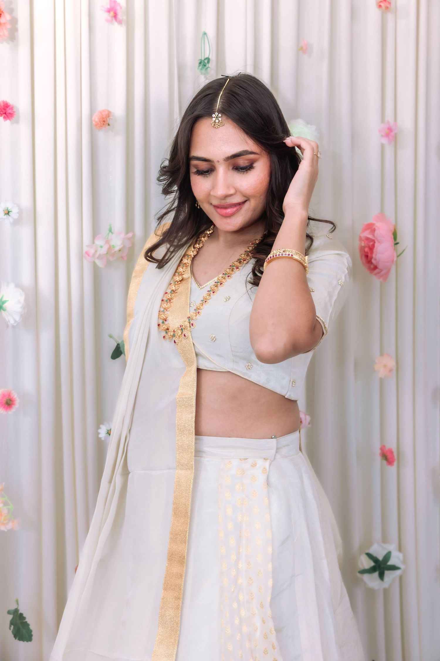Smiling woman in elegant white lehenga with gold jewelry and maang tikka at floral backdrop.