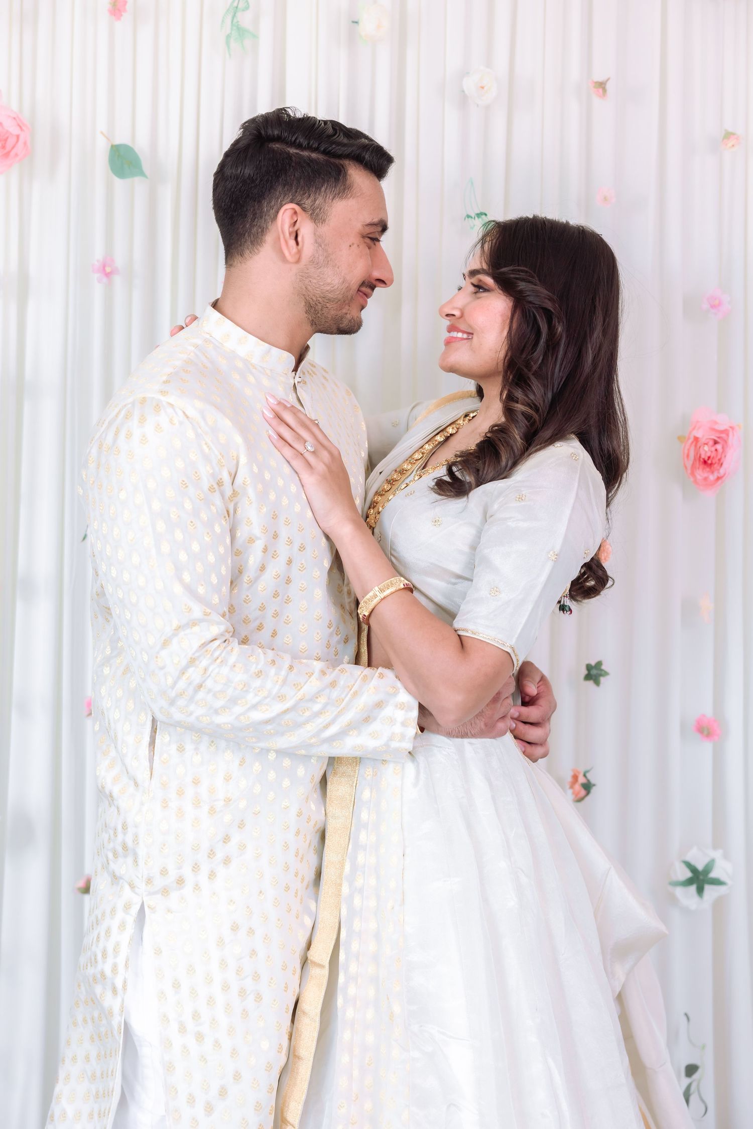 Indian couple in white traditional attire share a romantic gaze at a floral decorated celebration.