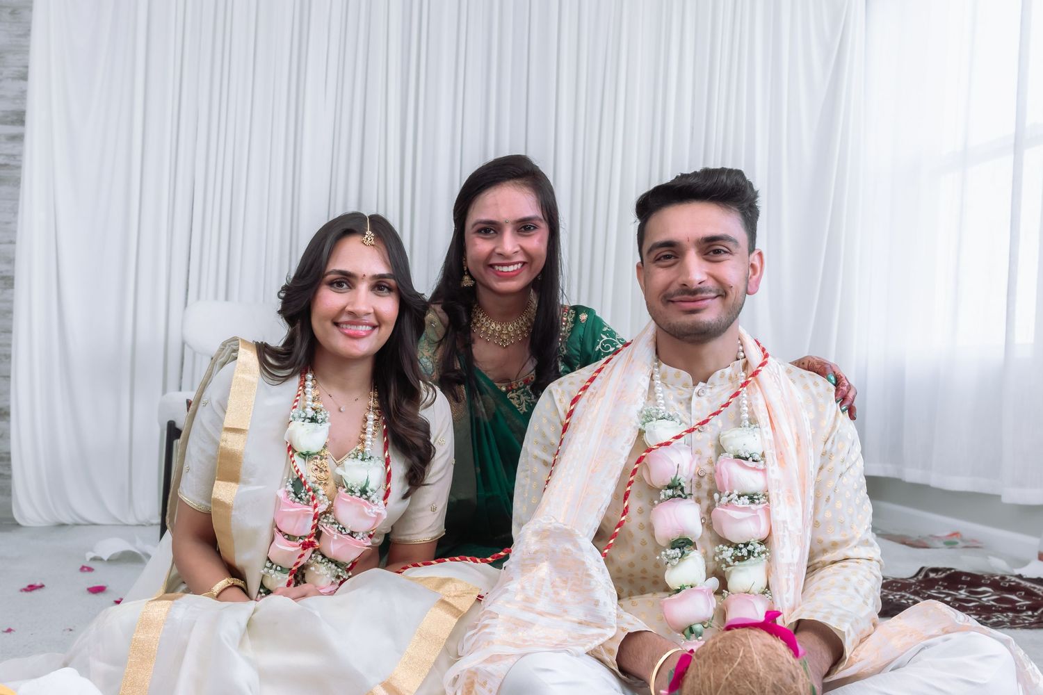 Indian bride and groom wearing floral garlands pose with a smiling female guest at their wedding.