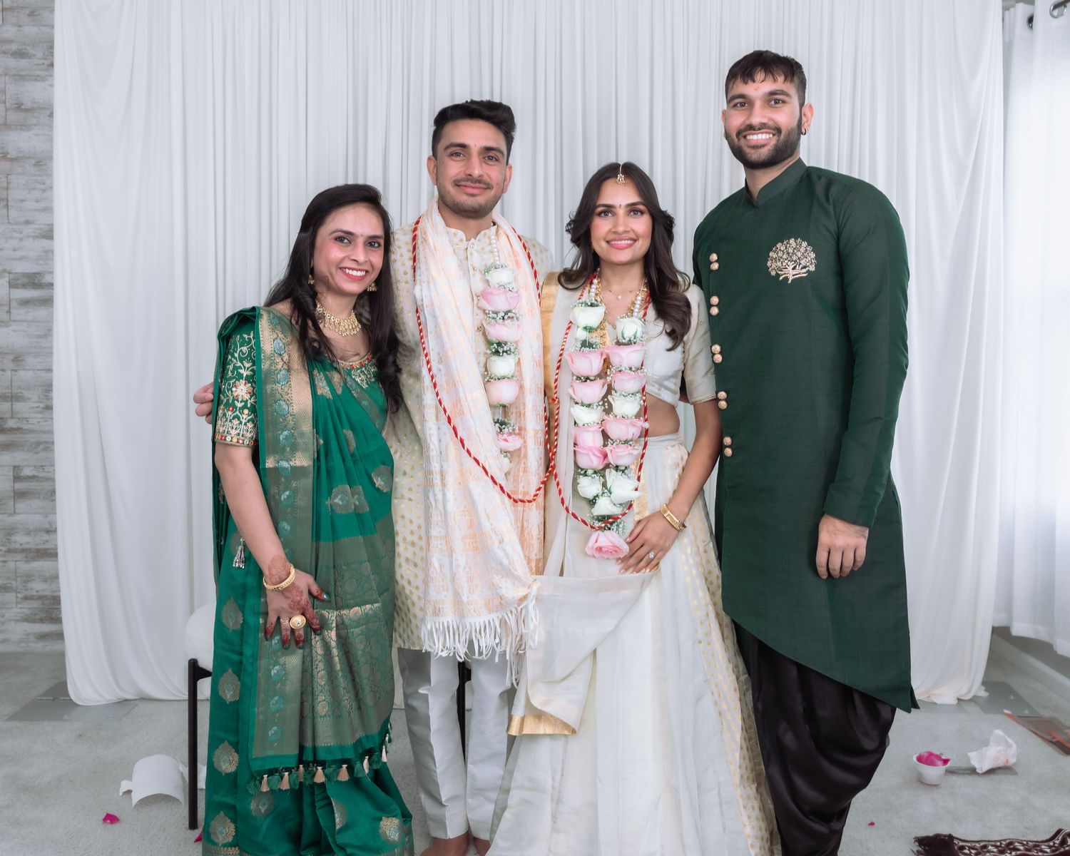 Indian wedding couple in floral garlands pose with family in traditional attire against white backdrop.