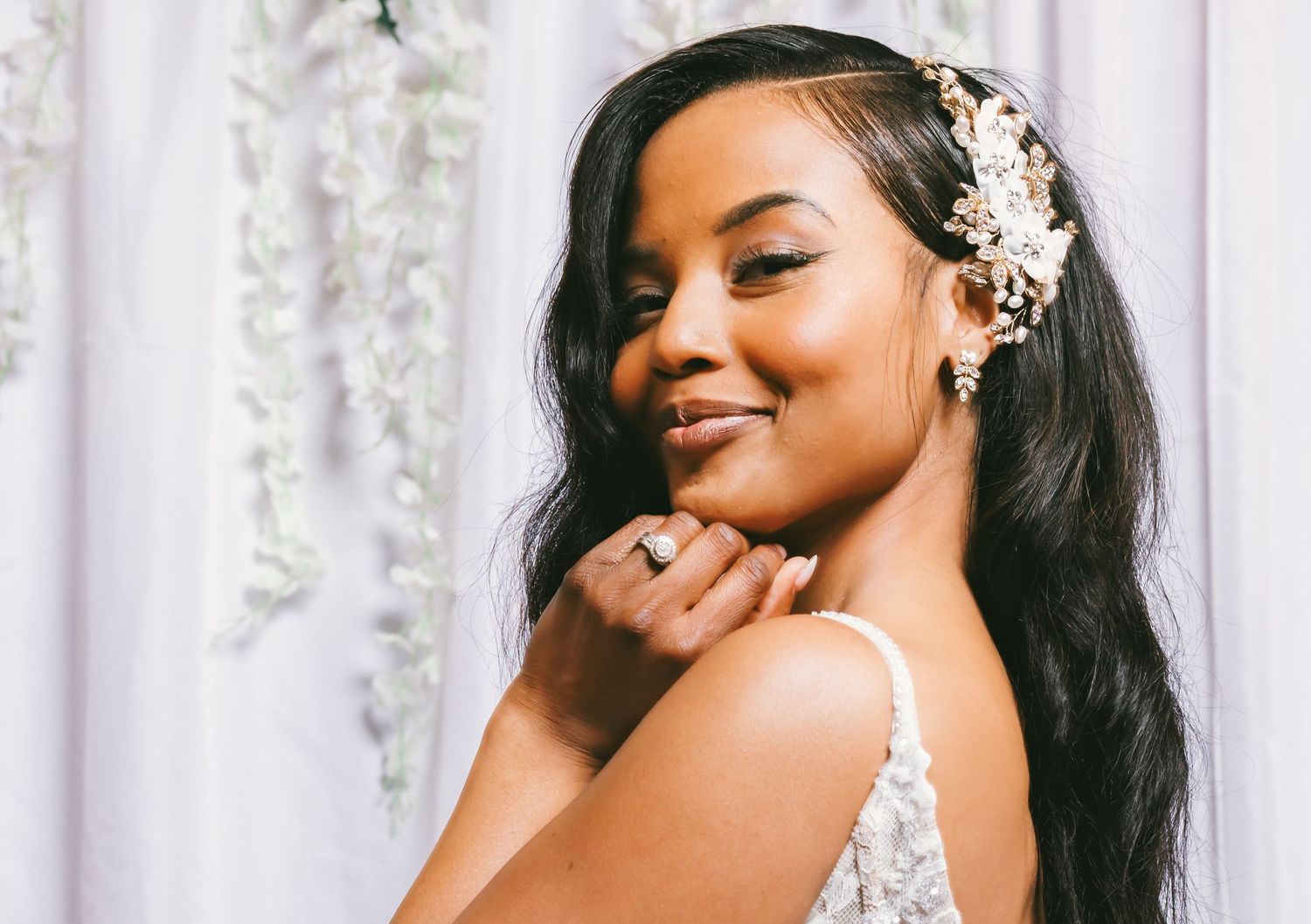 Stunning bride with pearl hair accessory in lace gown posing against white floral backdrop.