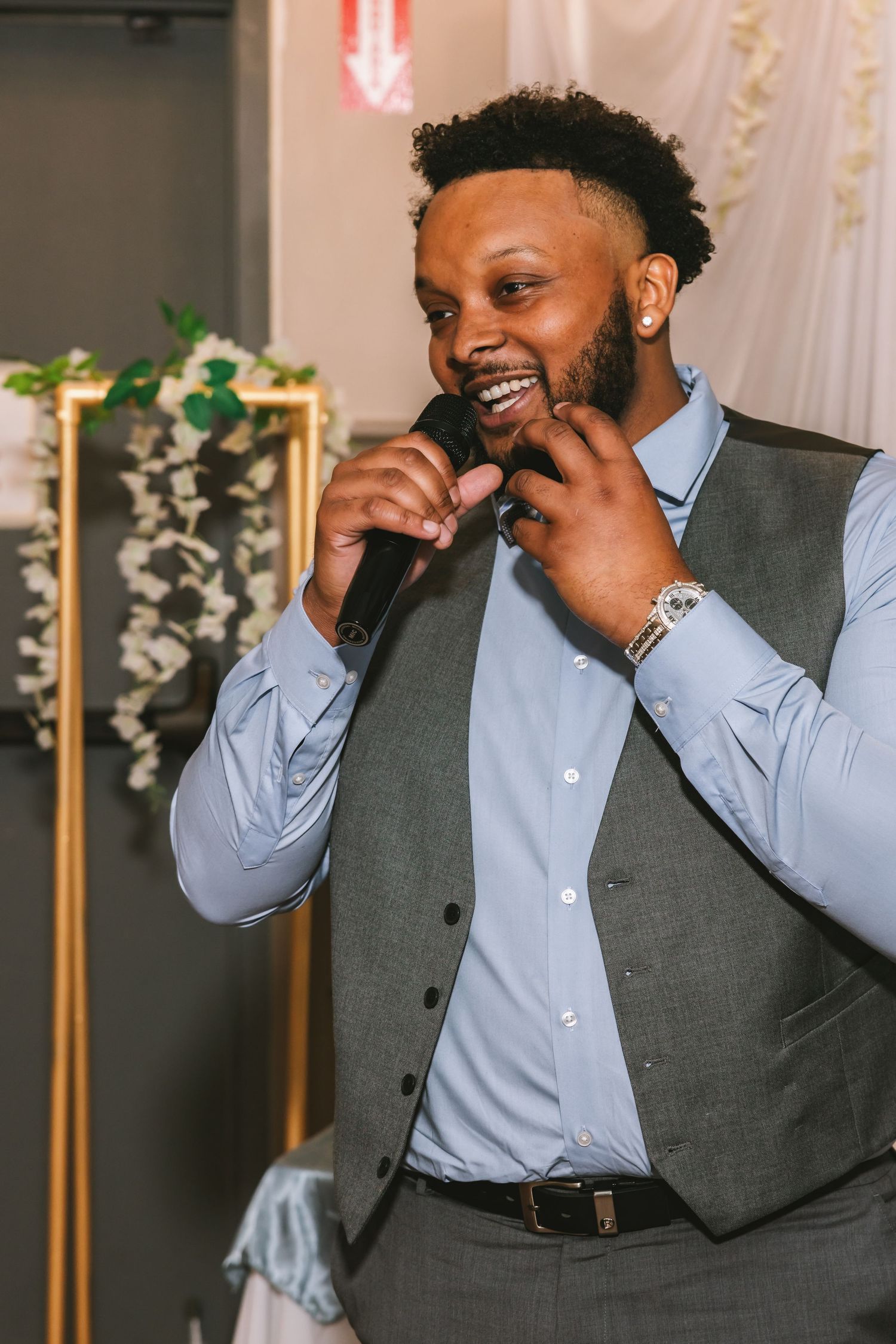 Man in gray vest and blue shirt smiles while speaking into microphone at a wedding reception.