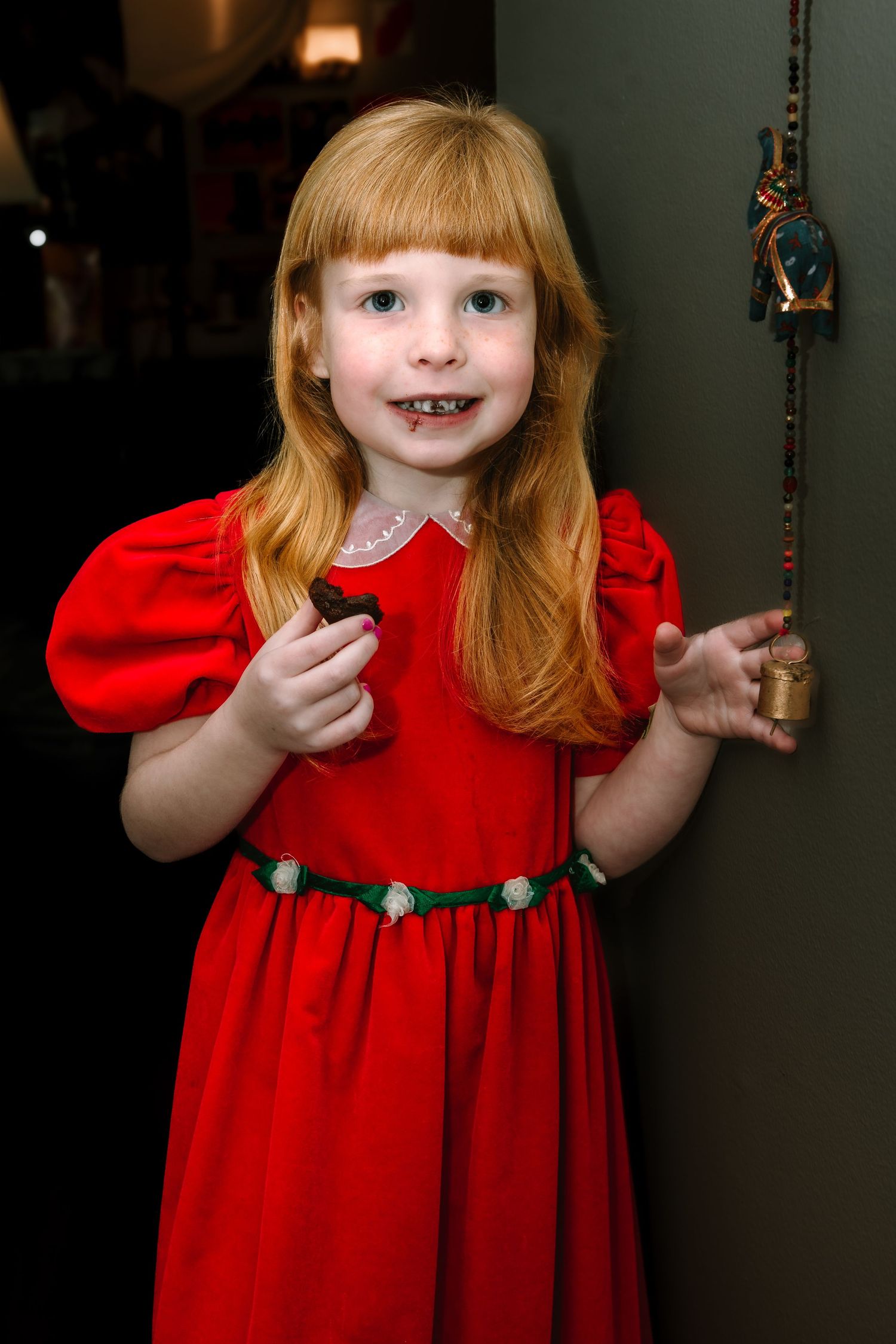Adorable young girl with red hair wearing a red dress smiling while holding a small object near a doorway.