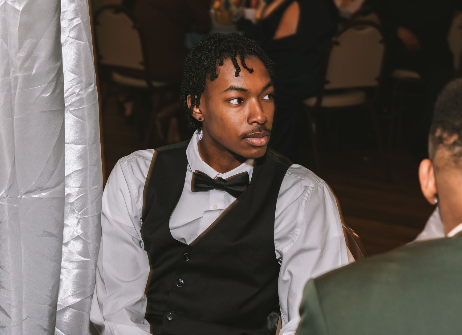 Young man in black vest and bow tie seated at a wedding reception.
