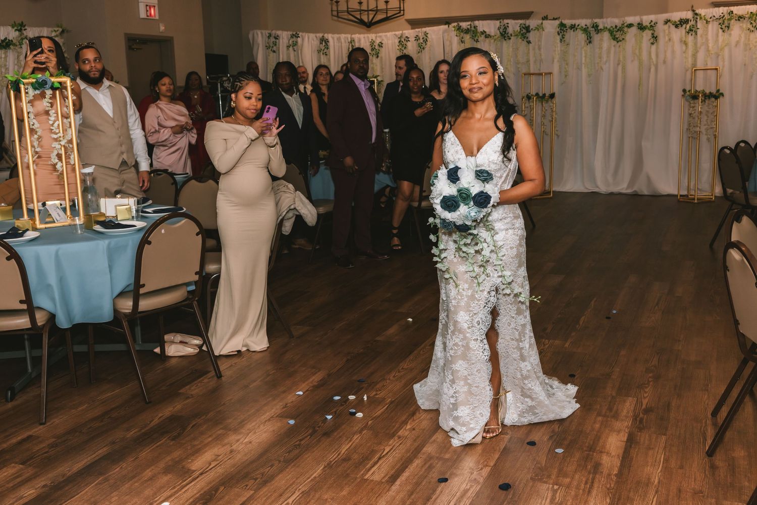 Bride walks down the aisle in a lace gown holding a blue and white bouquet at indoor wedding.
