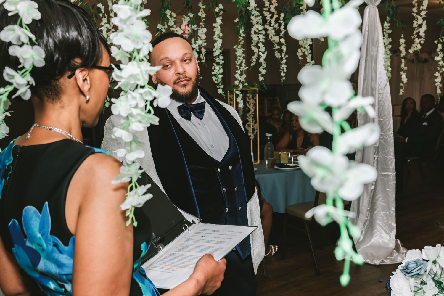 Groom in navy blue vest and bow tie listens attentively during wedding ceremony beneath floral arch.