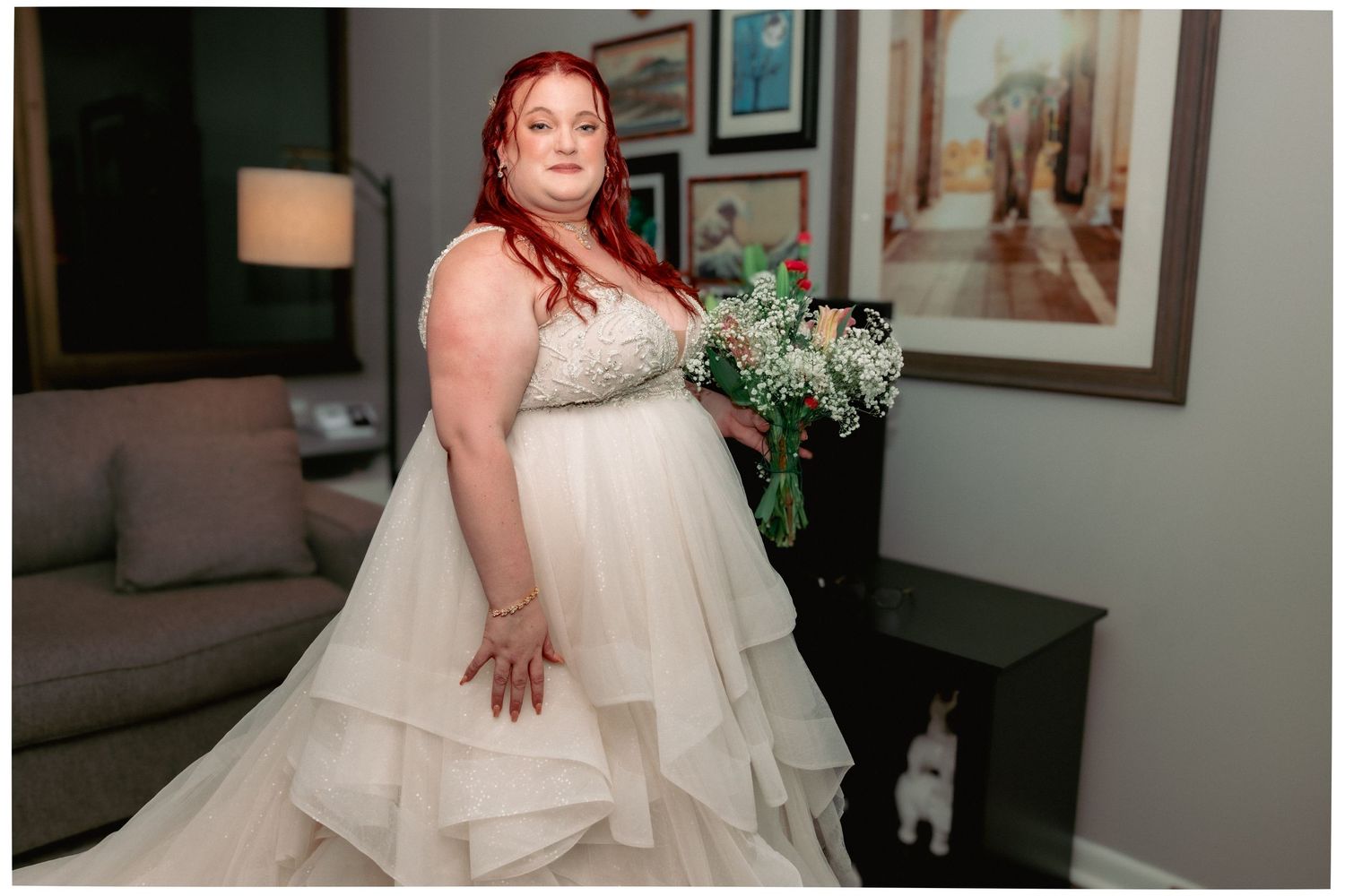 Red-haired bride in layered tulle wedding gown holds bouquet while posing in elegant hotel room.