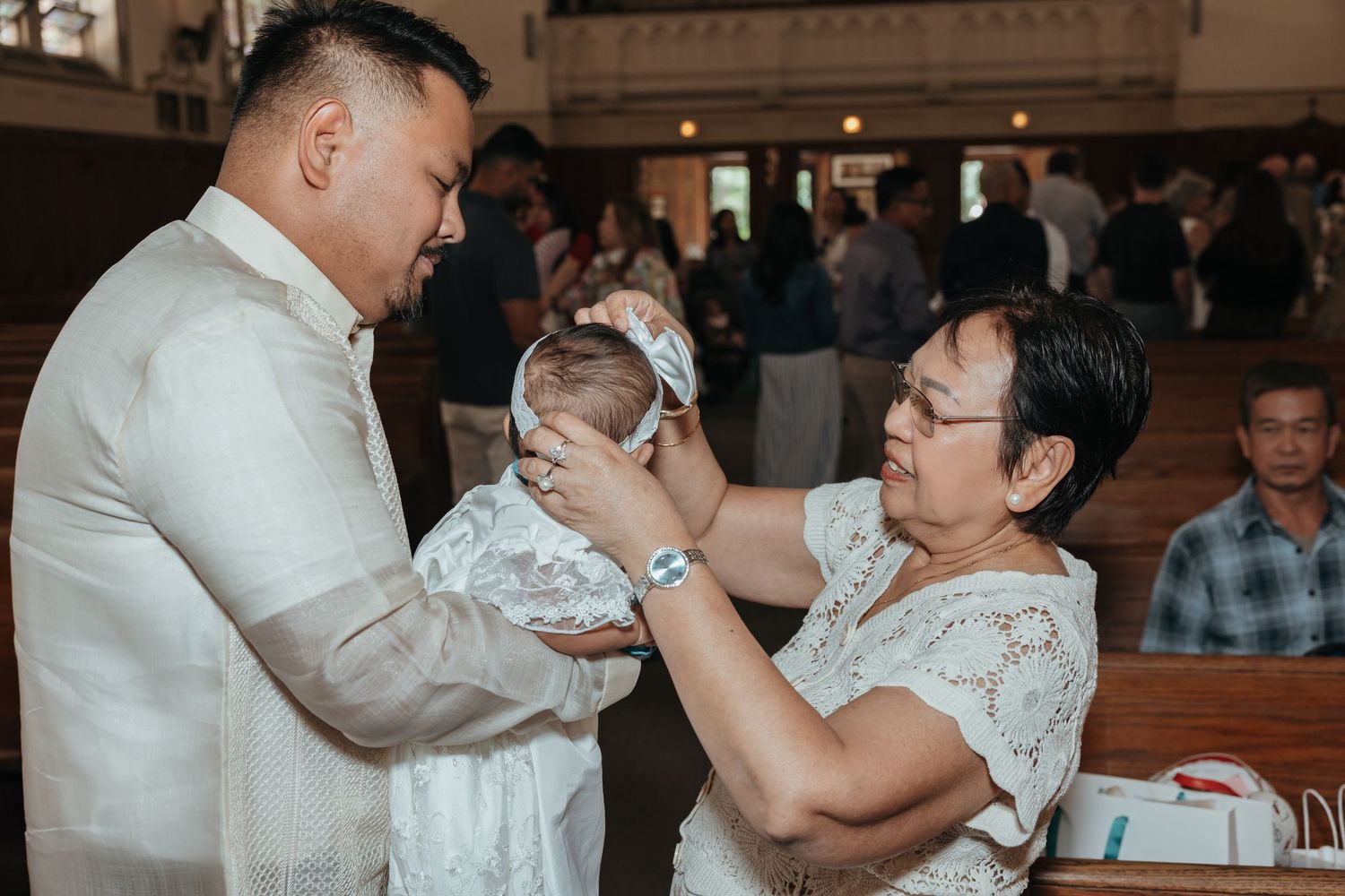 Man in barong receives baby in christening outfit from woman in church.