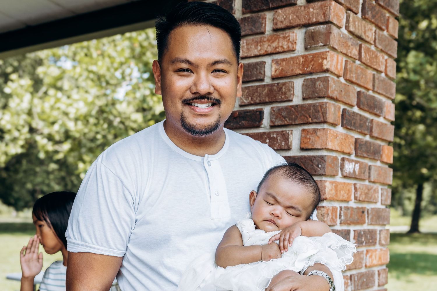 Smiling Asian father holds sleeping baby in white dress near brick wall outdoors.
