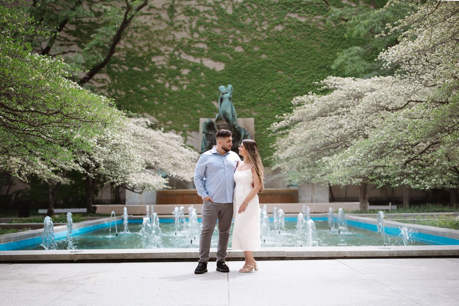 Couple standing by a fountain with blooming trees in a lush garden setting.