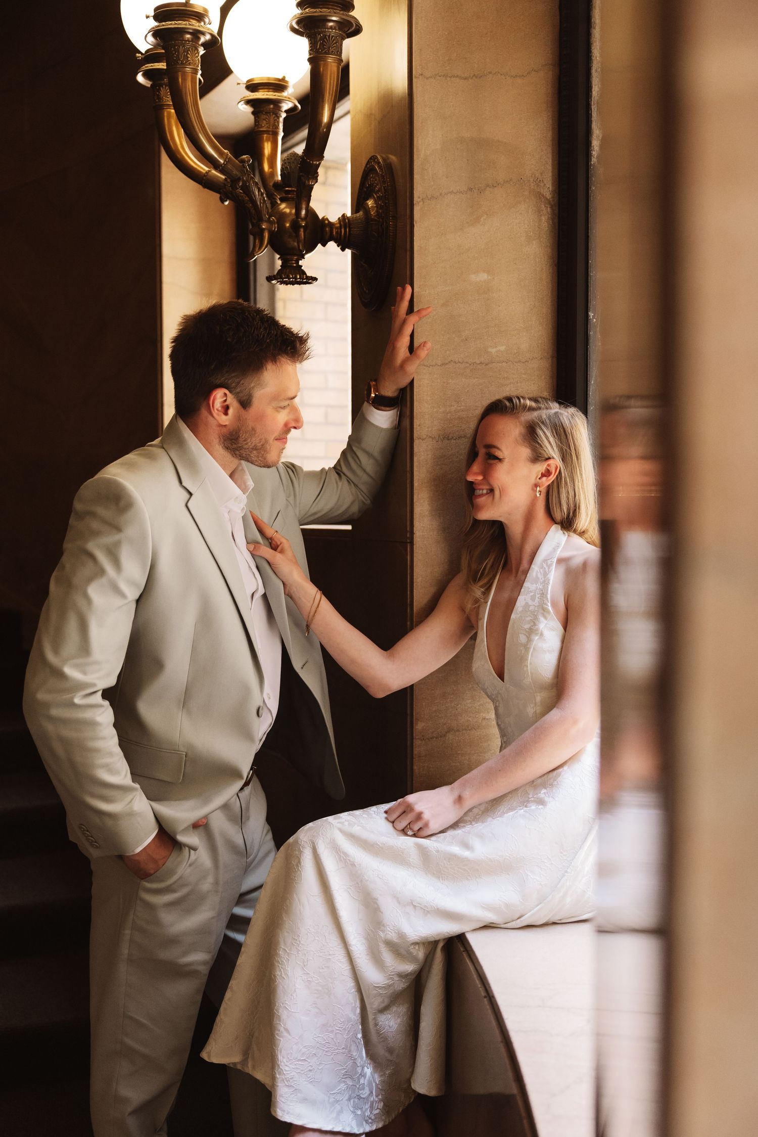 Bride in white halter gown and groom in sage suit share a joyful moment beside a golden chandelier.