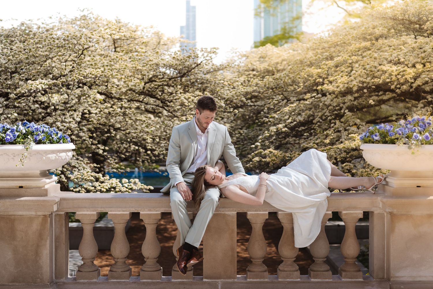 Bride reclines gracefully on a stone balustrade as groom gazes at her amid blooming white trees.