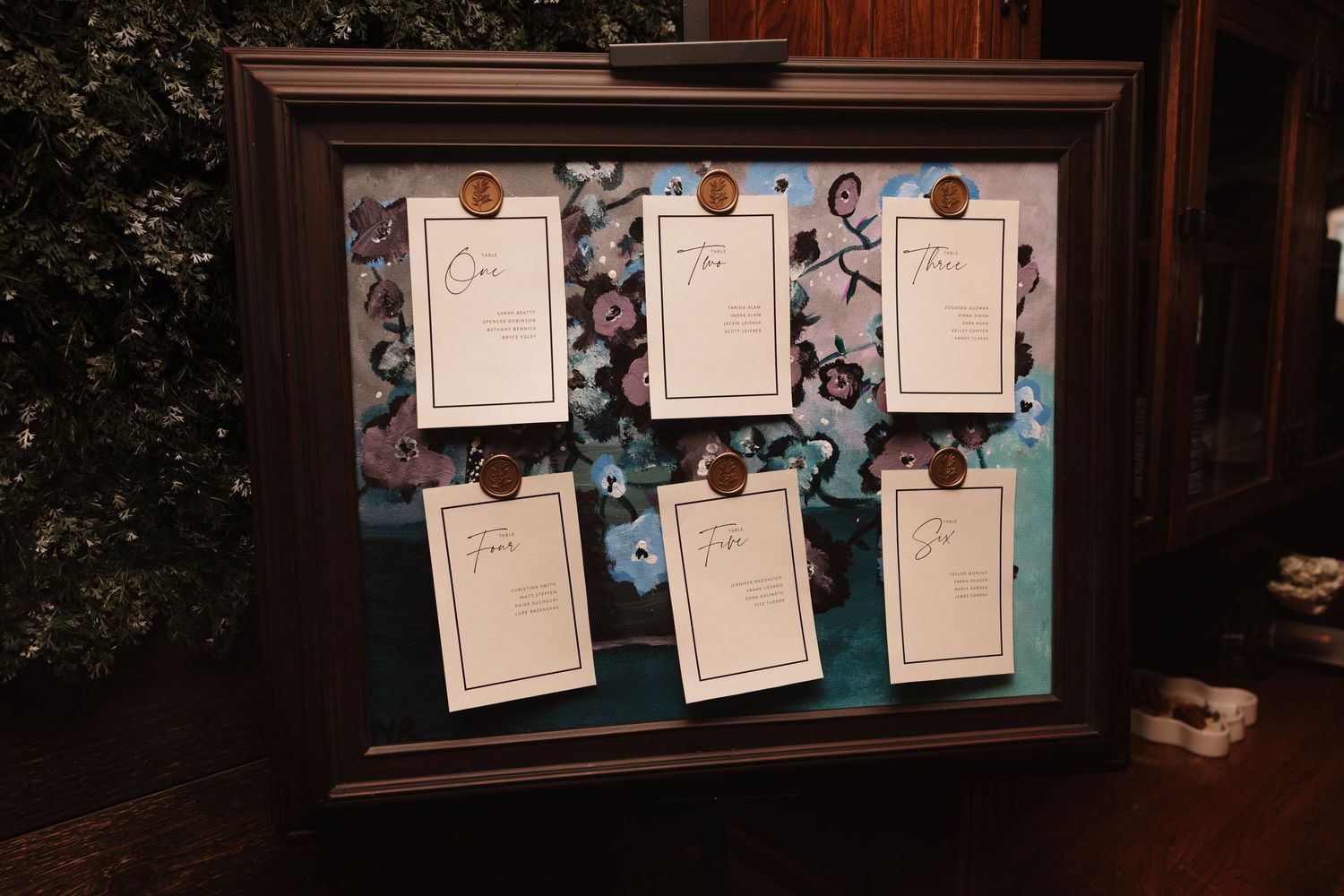 Elegant wedding seating chart display in dark frame with floral background and wax seal cards.