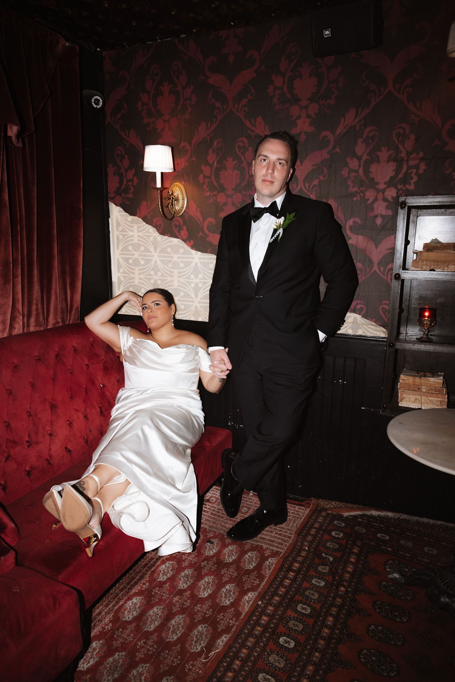Bride in white satin gown reclining on red velvet sofa while groom stands in dark luxe room.