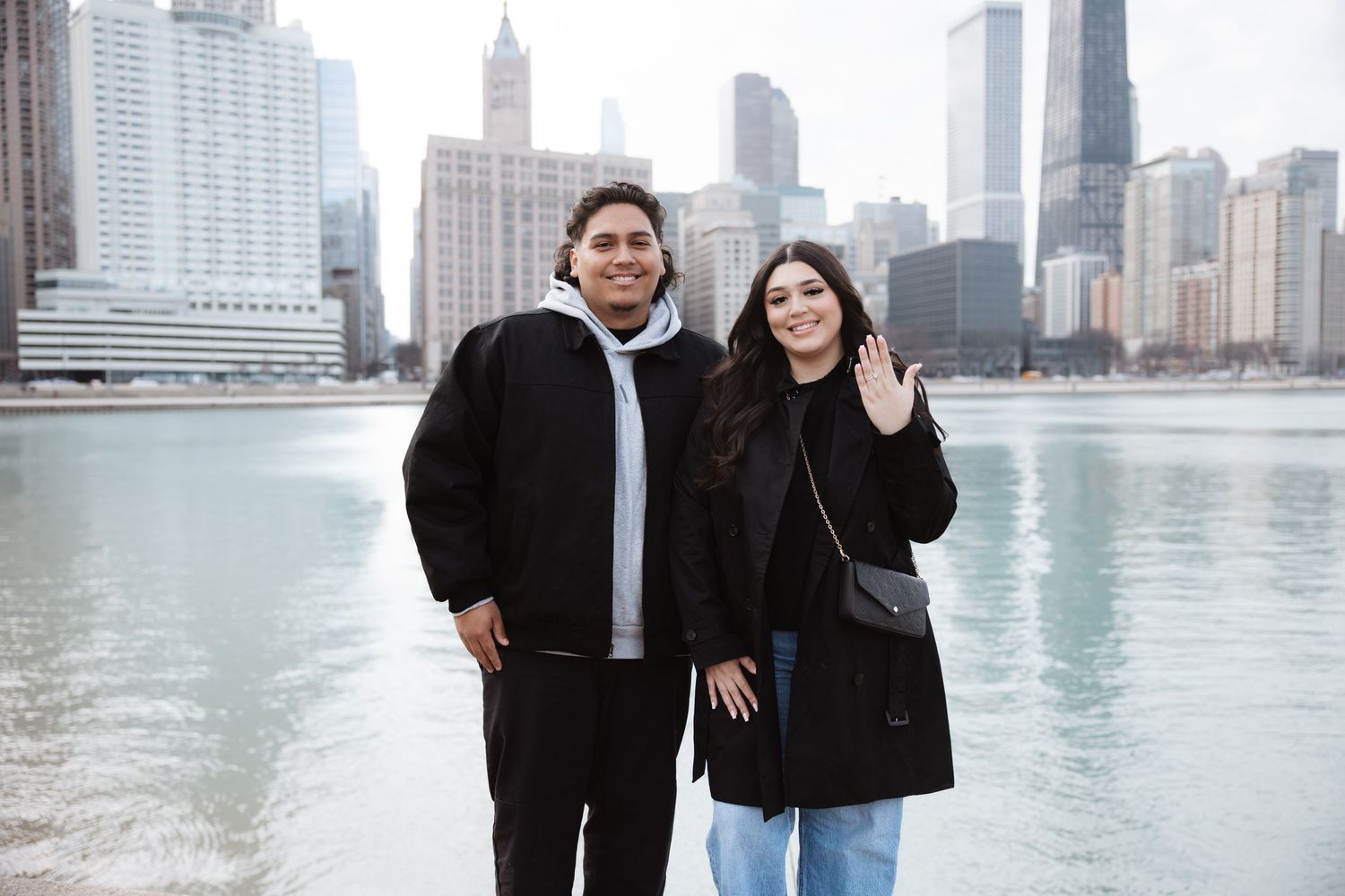 Engaged couple smiles together showing ring by Chicago River with downtown skyline backdrop.