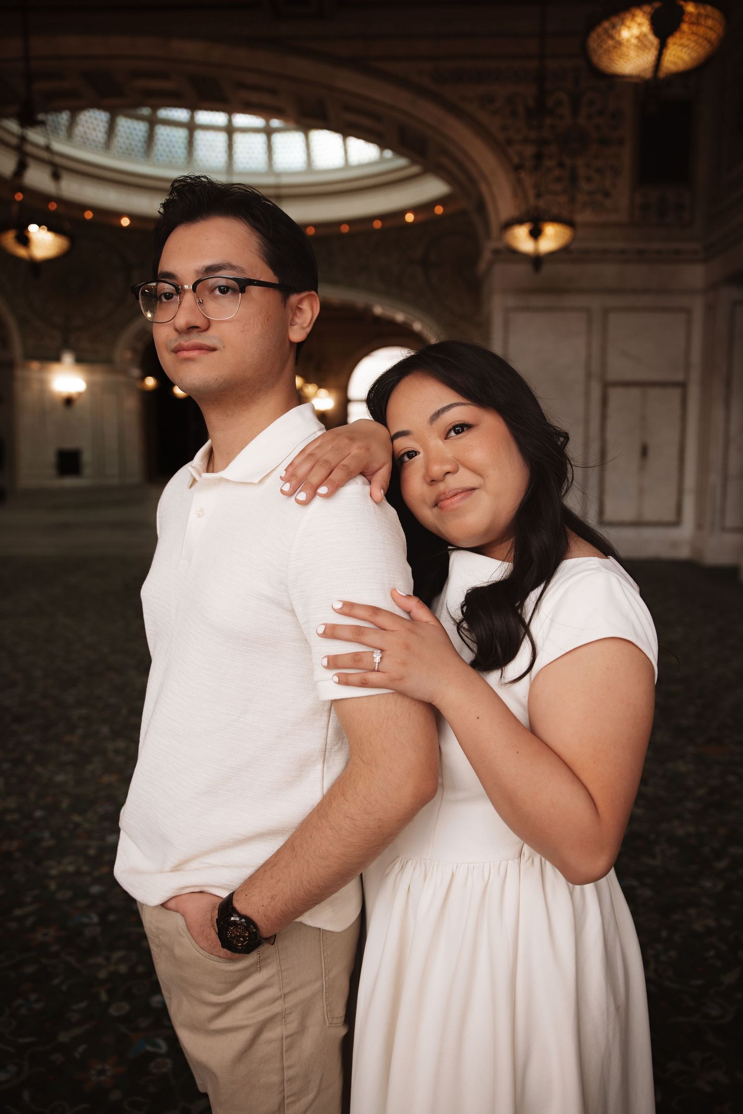 Couple in matching white outfits posing in grand architectural space, woman embracing man from behind.