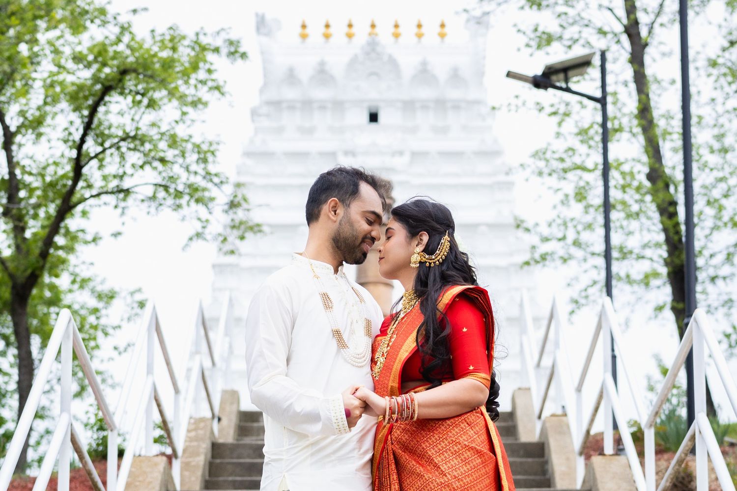Indian couple in traditional attire sharing a romantic moment before a white temple with lush green trees.