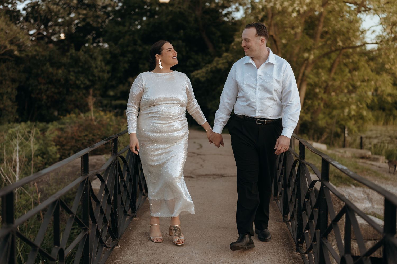 Couple holds hands and smiles while walking across a wooden bridge at golden hour.