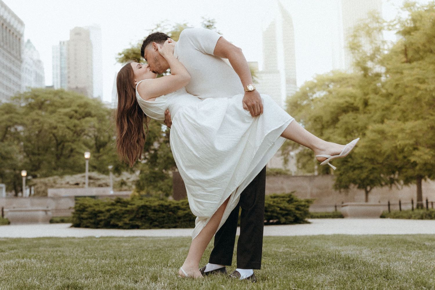 Couple sharing a romantic dip kiss in a city park, bride in white dress, groom in black suit, urban skyline backdrop.