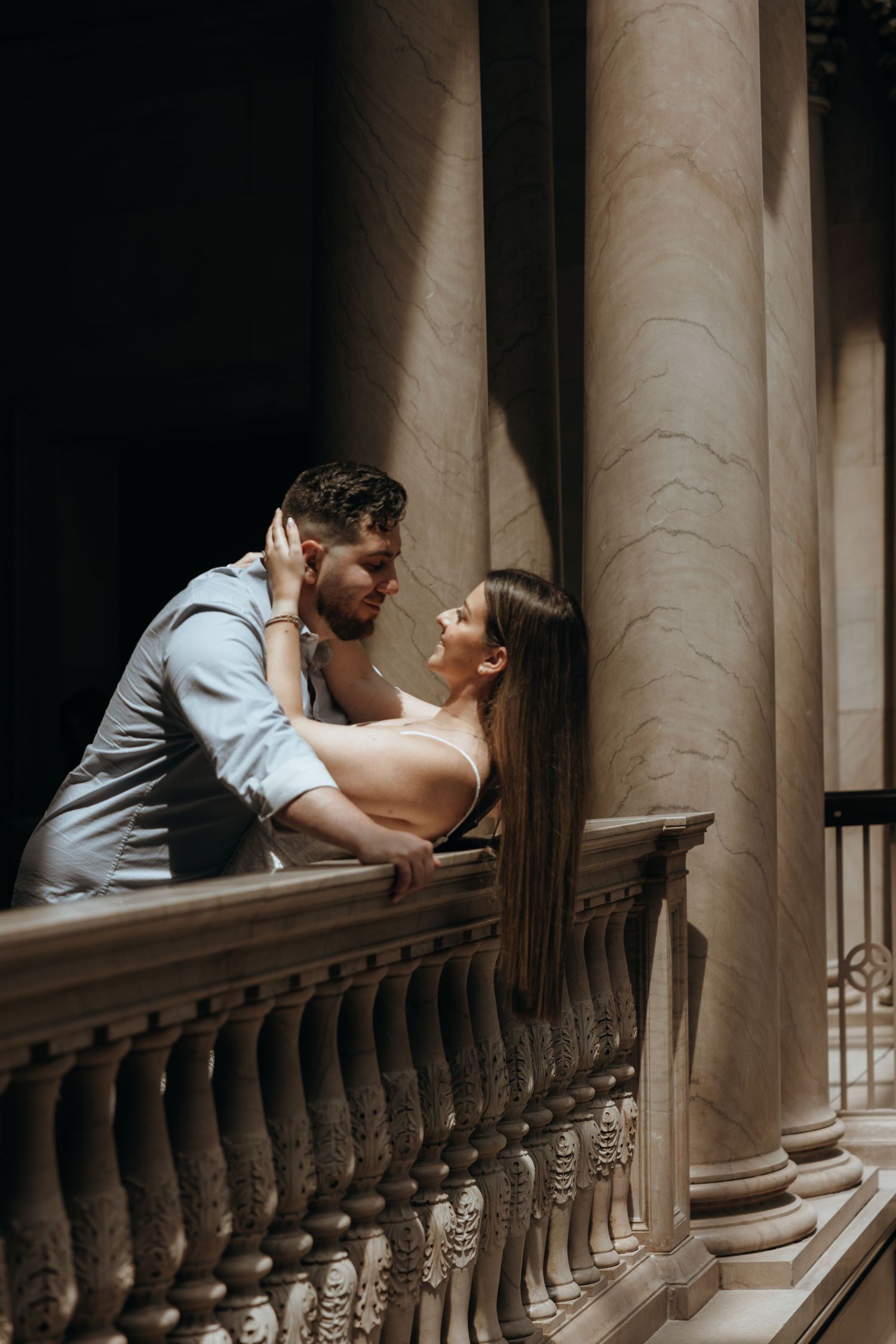 Couple leaning in for a kiss on a marble balcony with tall classical columns in dramatic lighting.